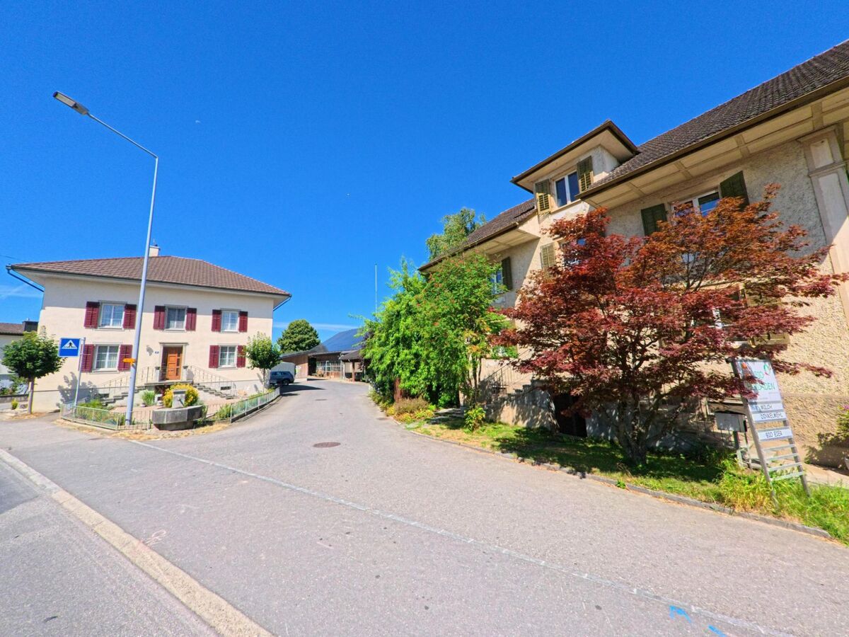 Zwei gepflegte Gebaeude mit Fensterlaeden und Vorgartenpraegung stehen in Buttisholz (6018, Kanton Luzern) an einer leicht ansteigenden Quartierstrasse unter blauem Himmel.