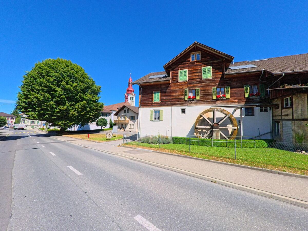 In Buttisholz (6018, Kanton Luzern) steht ein traditionelles Wohnhaus mit gruenen Fensterlaeden und grossem Wasserrad an der Strasse, im Hintergrund ist der rote Kirchturm sichtbar.