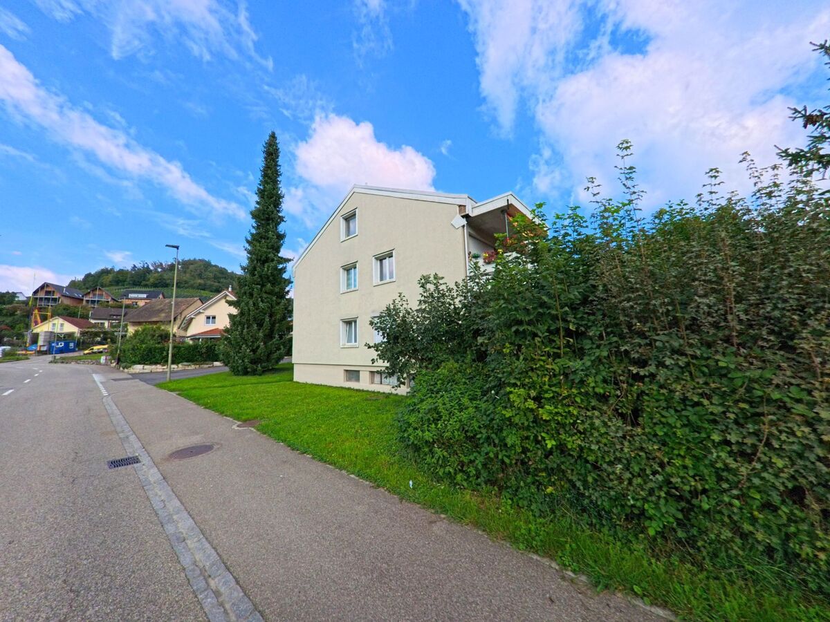 Entlang einer ruhigen Strasse in Buus (4463, Kanton Basel-Landschaft) steht ein schlichtes Mehrfamilienhaus neben dichter Vegetation und Blick auf huegelige Wohnlagen.