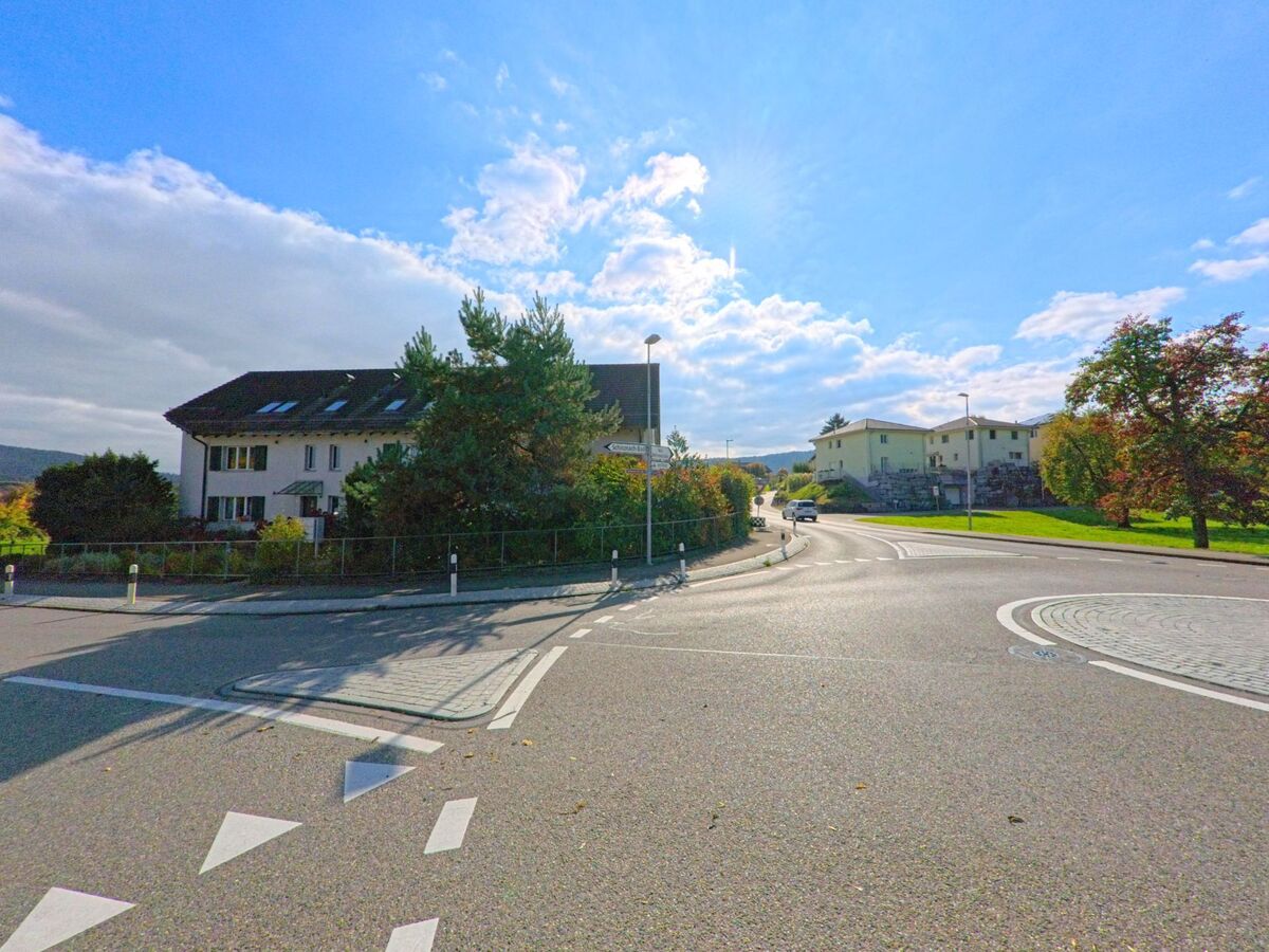 An einer breiten Kreuzung in Schinznach (5108, Kanton Aargau) stehen Wohnhäuser mit gepflegten Gärten, im Hintergrund führt die Strasse durch eine grüne Landschaft.