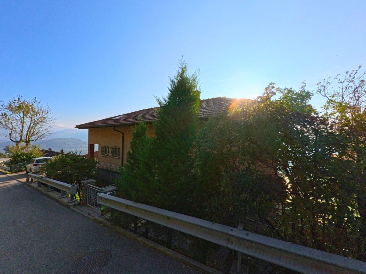 Zwischen Baeumen und Hecken liegt in Cademario (6936, Kanton Tessin) ein Wohnhaus an einer Hangstrasse, waehrend die Sonne tief ueber der Landschaft steht.