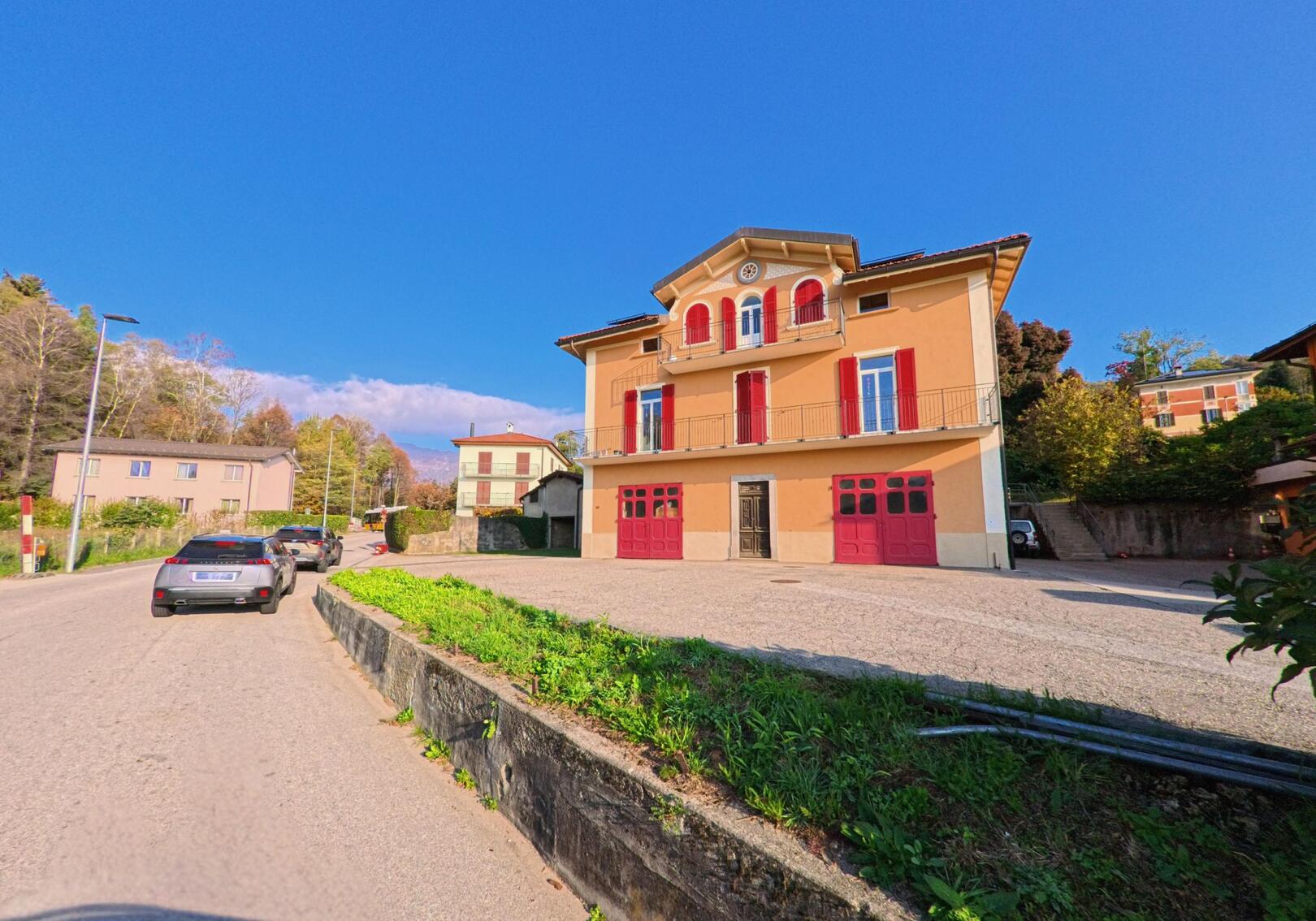 An einer ruhigen Strasse in Cademario (6936, Kanton Tessin) steht ein markantes Gebäude mit roten Fensterlaeden und freiem Vorplatz unter klarem Himmel.