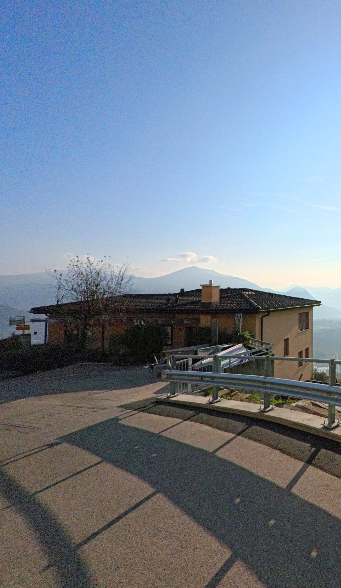 An einer Wegbiegung mit Geländer öffnet sich in Cademario (6936, Kanton Tessin) der Blick über ein Wohnhaus hinweg auf die ruhige Hügellandschaft.