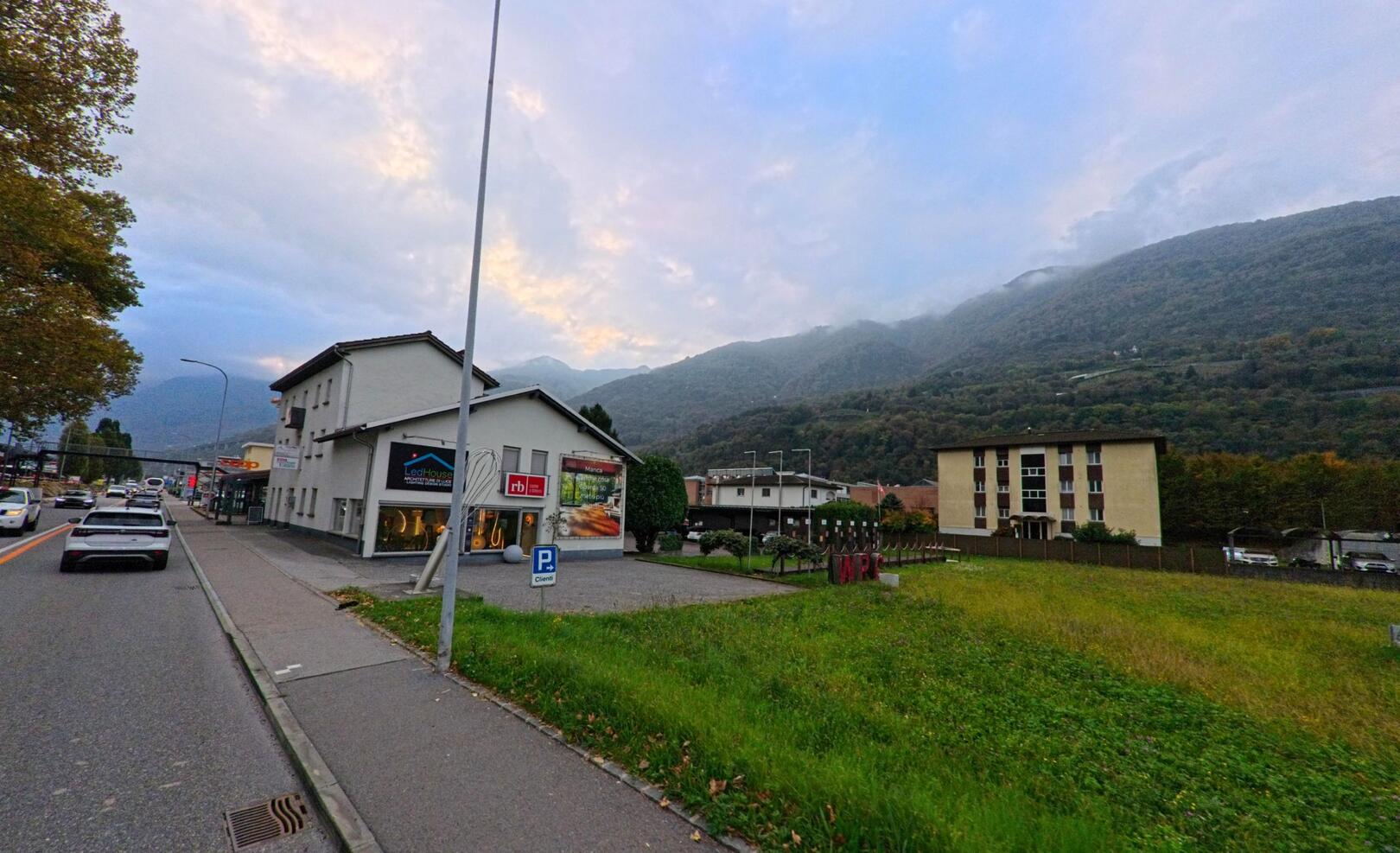 Entlang der Hauptstrasse in Cadenazzo (6593, Kanton Tessin) stehen moderne Gebäude vor der Kulisse bewaldeter Berghänge unter einem bewölkten Himmel.