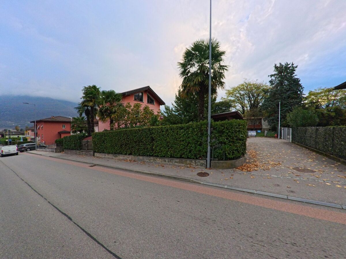 Ein rosafarbenes Wohnhaus mit Palmen und dichter Hecke steht an einer ruhigen Strasse in Cadenazzo (6593, Kanton Tessin), mit Blick auf bewaldete Hügel im Hintergrund.