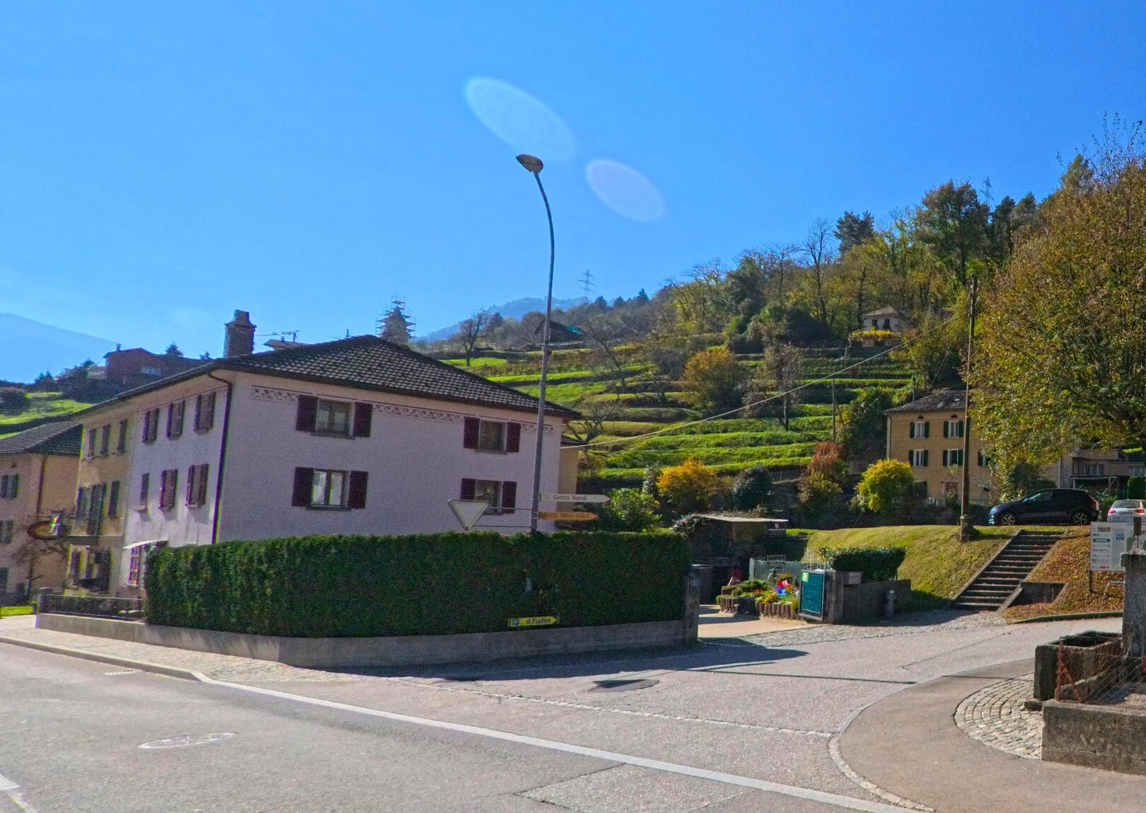 An einer ruhigen Kreuzung mit Wohnhaeusern und terrassierten Haengen zeigt sich das Dorfbild von Cama (6557, Kanton Graubuenden) bei klarem Himmel.