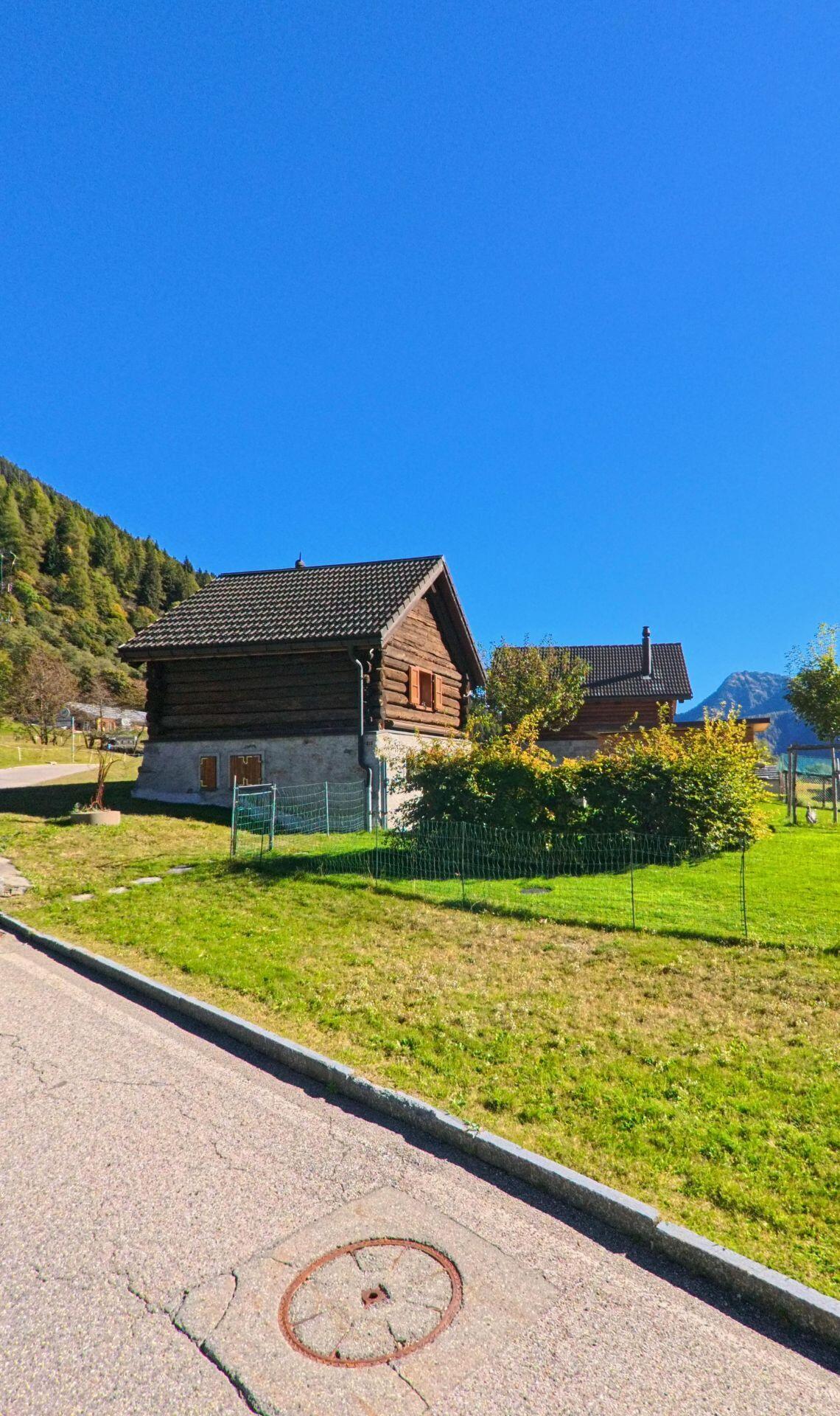 Ein kleines Holzhaus mit gemauertem Sockel steht in Campo (6684, Kanton Tessin) an einer ruhigen Strasse, umgeben von grüner Wiese und Bäumen.