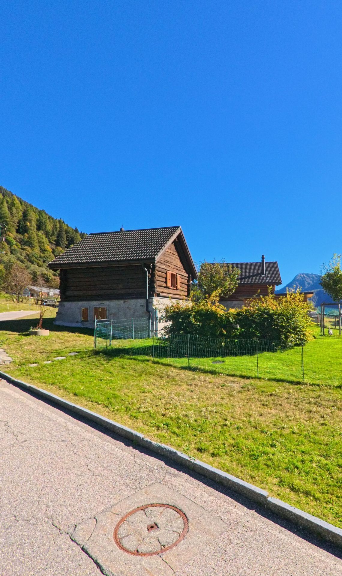 Ein kleines Holzhaus mit gemauertem Sockel steht in Campo (6684, Kanton Tessin) an einer ruhigen Strasse, umgeben von grüner Wiese und Bäumen.