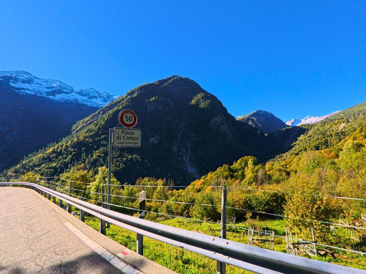Am Ortseingang von Campo (6684, Kanton Tessin) zeigt ein Schild „Piano di Campo“ den Beginn des Dorfgebiets an, umgeben von grünen Bergen und schneebedeckten Gipfeln.