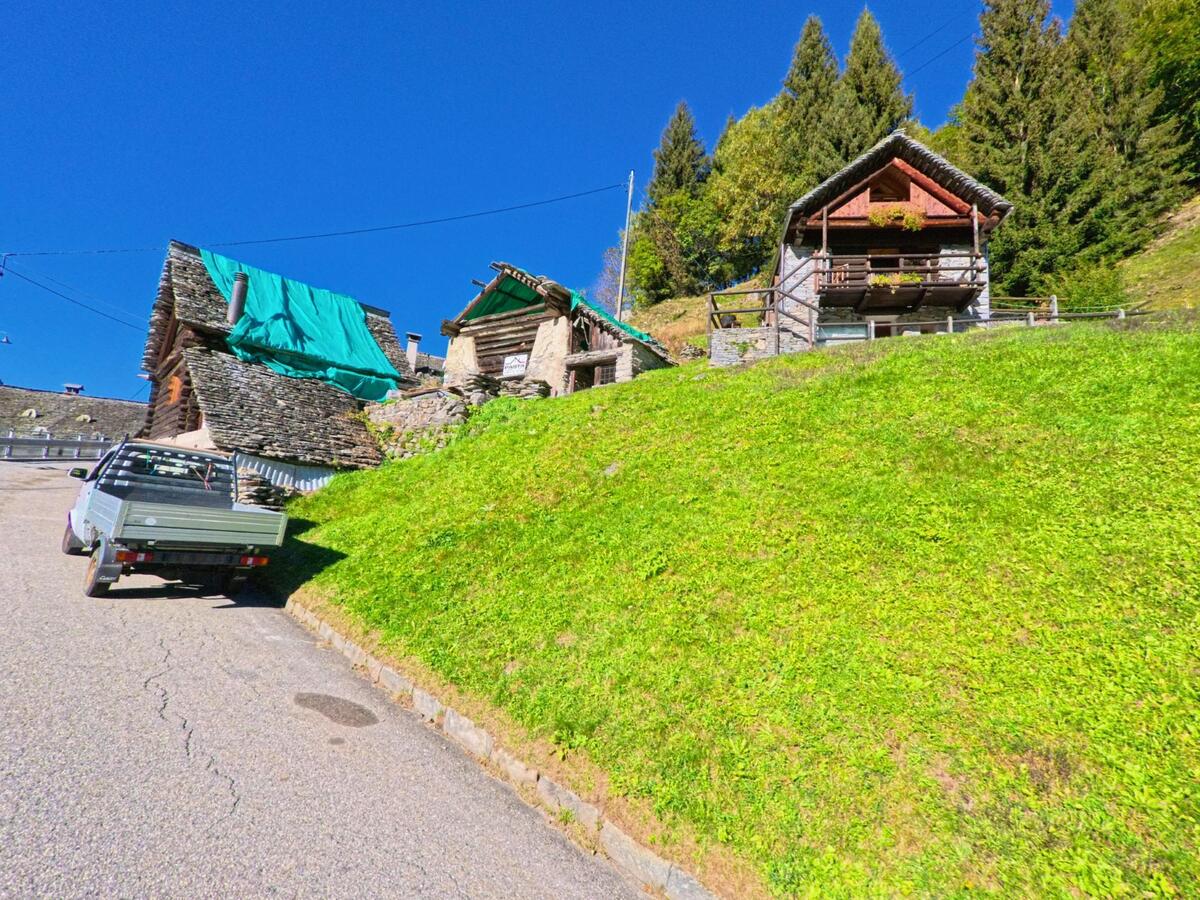 An einem steilen Hang in Campo (6684, Kanton Tessin) stehen traditionelle Steinhäuser mit Schieferdächern und Holzbalkonen, teils von grünen Planen bedeckt.