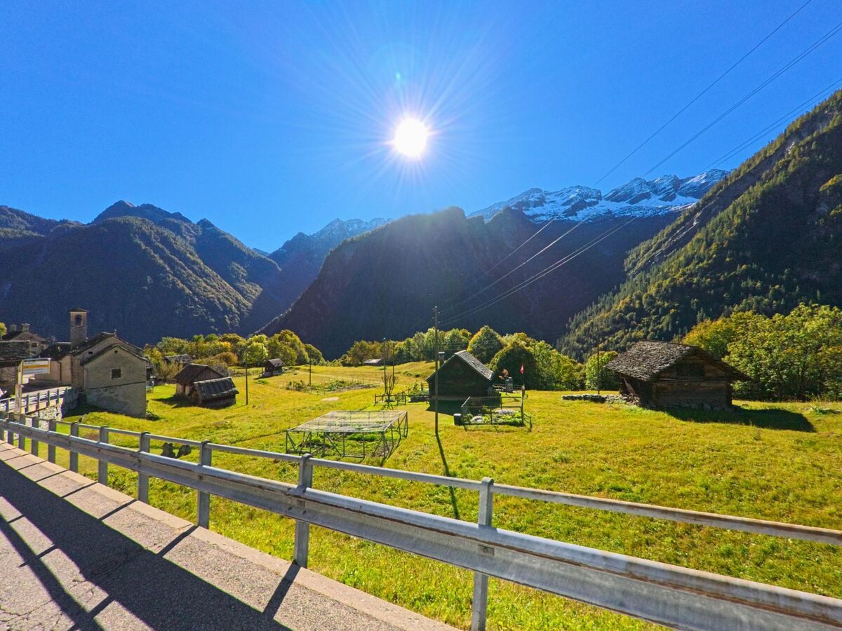 Bei strahlendem Sonnenschein breitet sich in Campo (6684, Kanton Tessin) eine weite Wiese mit kleinen Holzhütten aus, umgeben von hohen Bergen mit schneebedeckten Gipfeln.