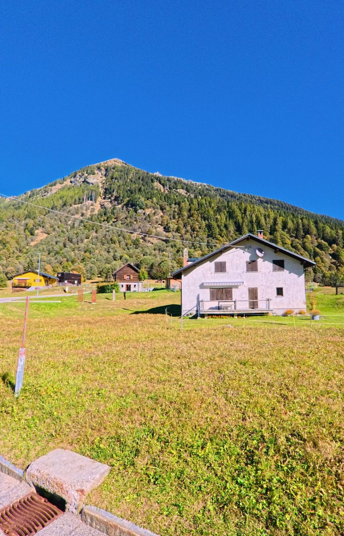 Ein helles Wohnhaus steht in Campo (6684, Kanton Tessin) vor einem bewaldeten Berghang unter klarem, blauem Himmel in ruhiger Lage.