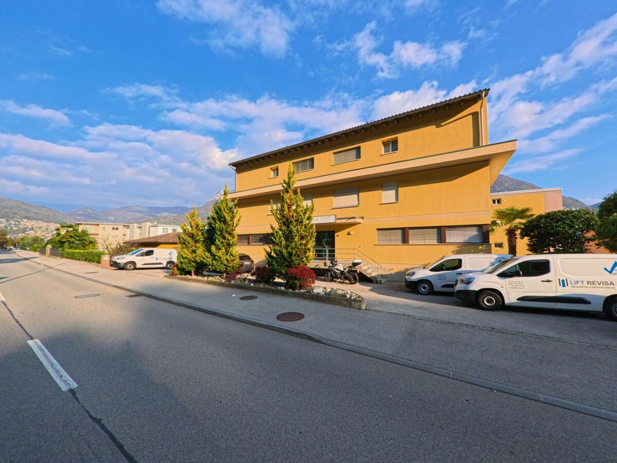 Ein groesseres Gebaeude mit Vorplatz und parkierenden Fahrzeugen liegt an einer ruhigen Strasse in Canobbio (6952, Kanton Tessin) vor sanften Huegeln.