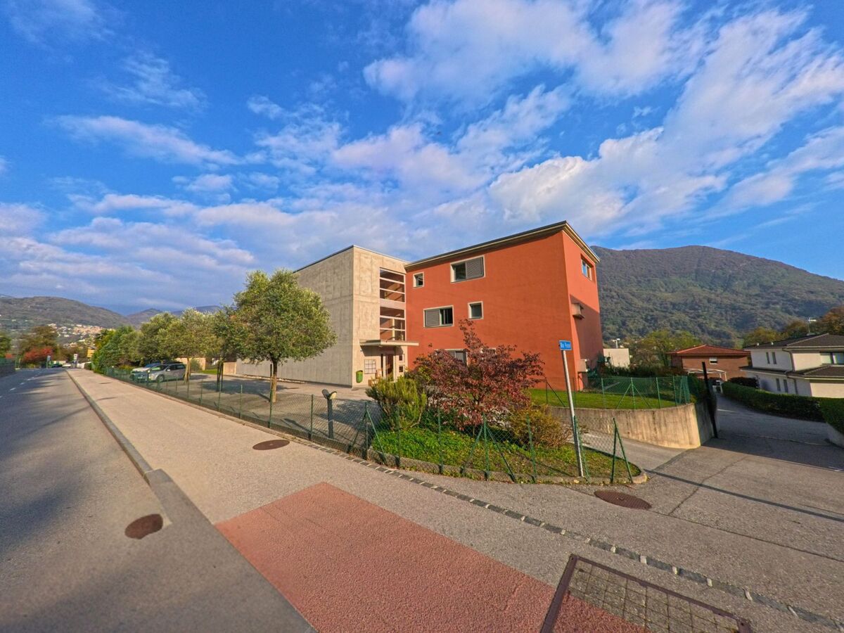 Ein modernes Mehrfamilienhaus mit Gehweg und Gruenflaechen praegt eine ruhige Strassenszene in Canobbio (6952, Kanton Tessin) vor bewaldeten Huegeln.