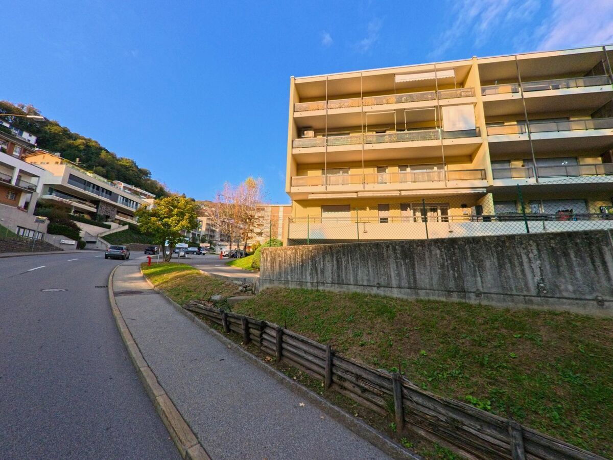 Entlang einer leicht ansteigenden Quartierstrasse mit Mehrfamilienhaeusern und Balkonen zeigt sich ein Wohngebiet in Canobbio (6952, Kanton Tessin) am Hang.