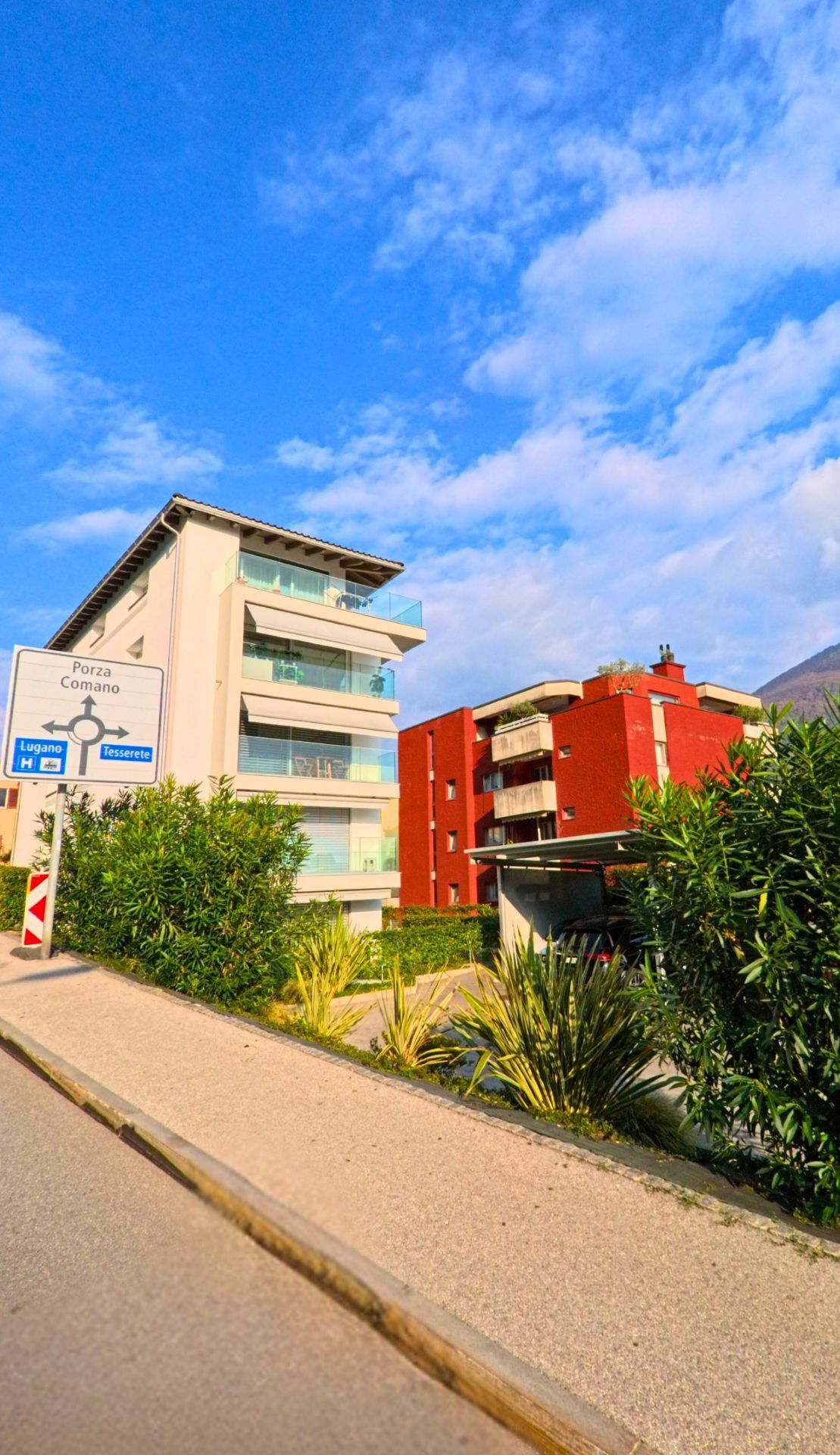 Moderne Wohnbauten mit Balkonen und Begruenung stehen an einer Strasse in Canobbio (6952, Kanton Tessin), begleitet von Wegweisern und freiem Himmel.