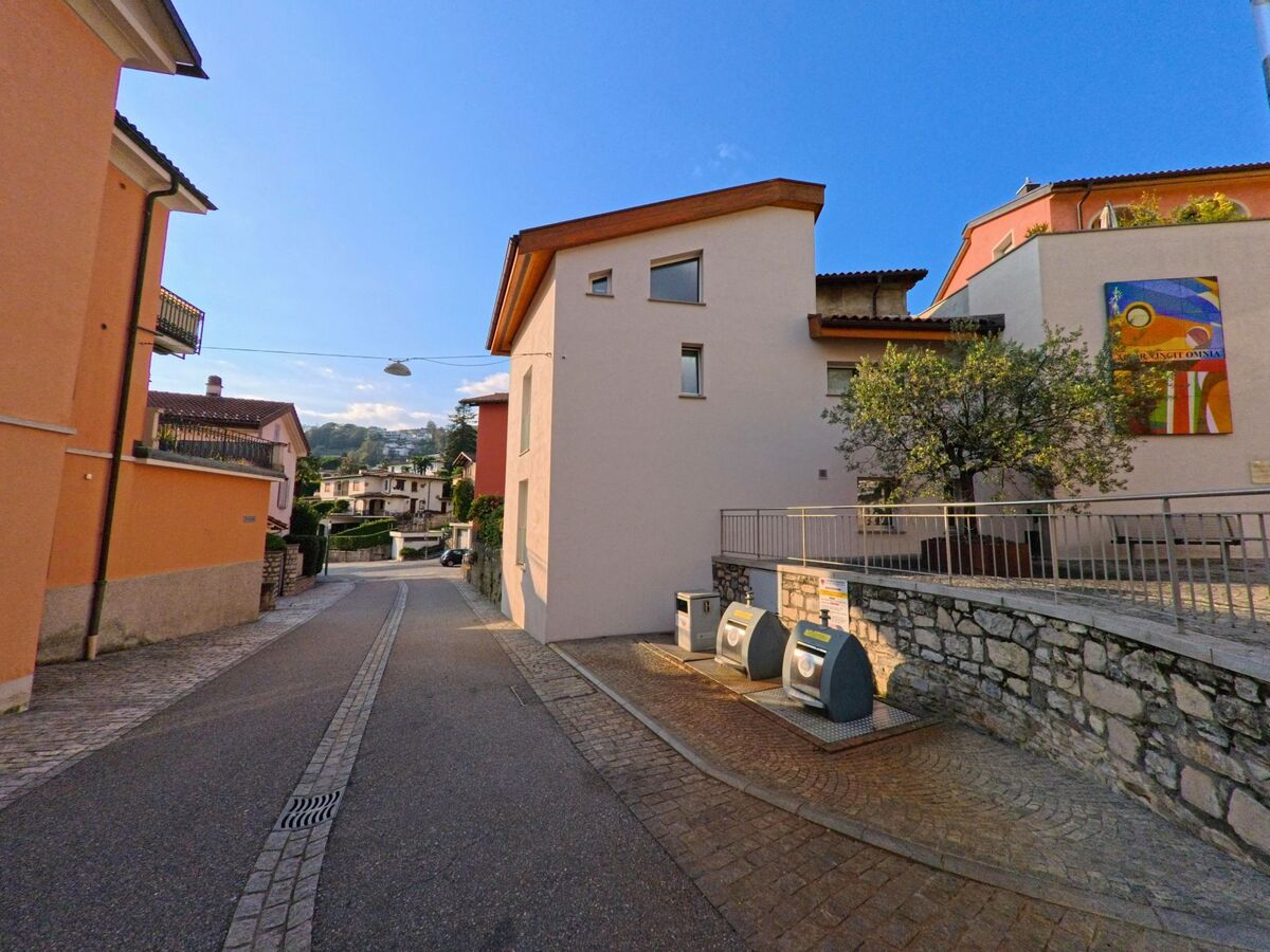 Eine schmale Gasse mit Wohnhaeusern und Sammelbehaeltern verlaeuft durch ein ruhiges Quartier in Canobbio (6952, Kanton Tessin) bei klarem Himmel.