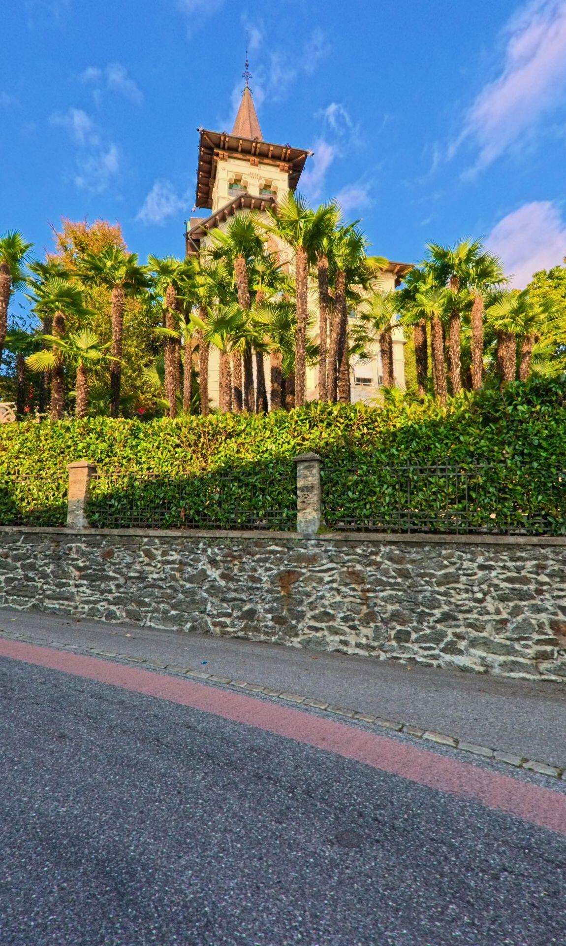 Hinter einer Steinmauer und Palmen erhebt sich ein turmartiges historisches Gebäude entlang der Strasse in Capriasca (6950, Kanton Tessin) bei sonnigem Wetter.