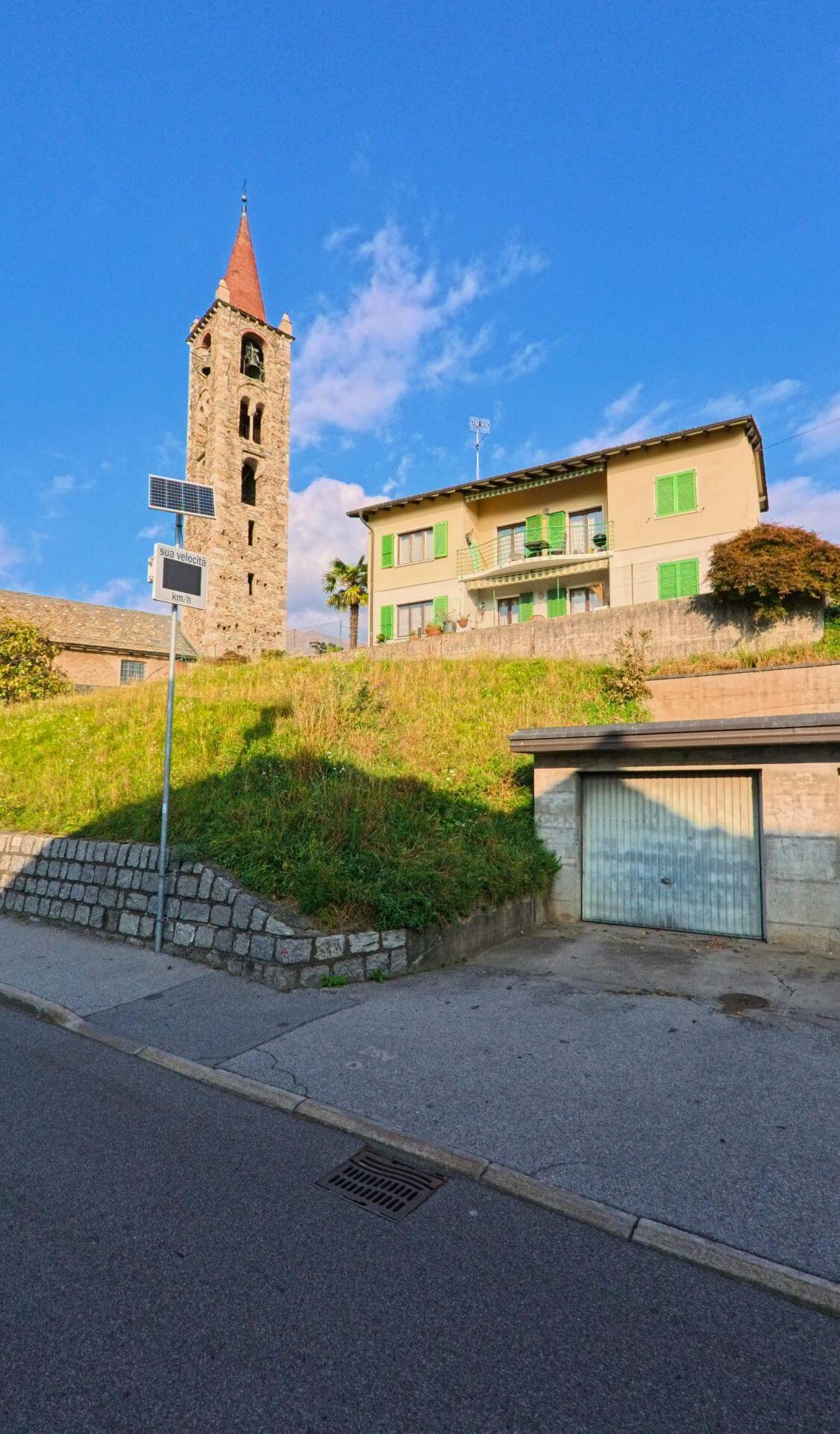Ein markanter Kirchturm aus Naturstein ragt neben Wohnhäusern an einem Hang über der Strasse in Capriasca (6950, Kanton Tessin) in den blauen Himmel.