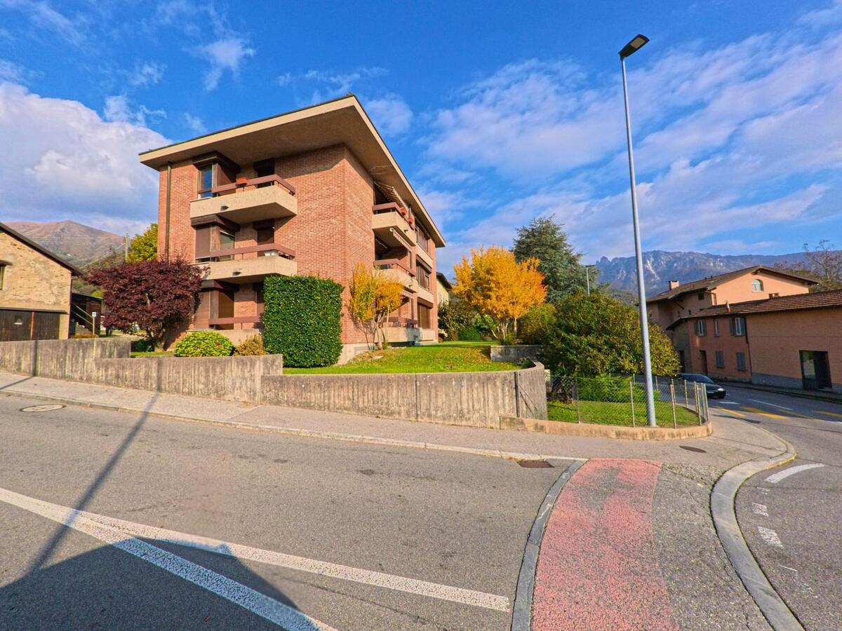 Ein Mehrfamilienhaus mit Balkonen und herbstlich gefärbten Bäumen steht an einer Strassenecke in Capriasca (6950, Kanton Tessin) vor bergiger Kulisse.