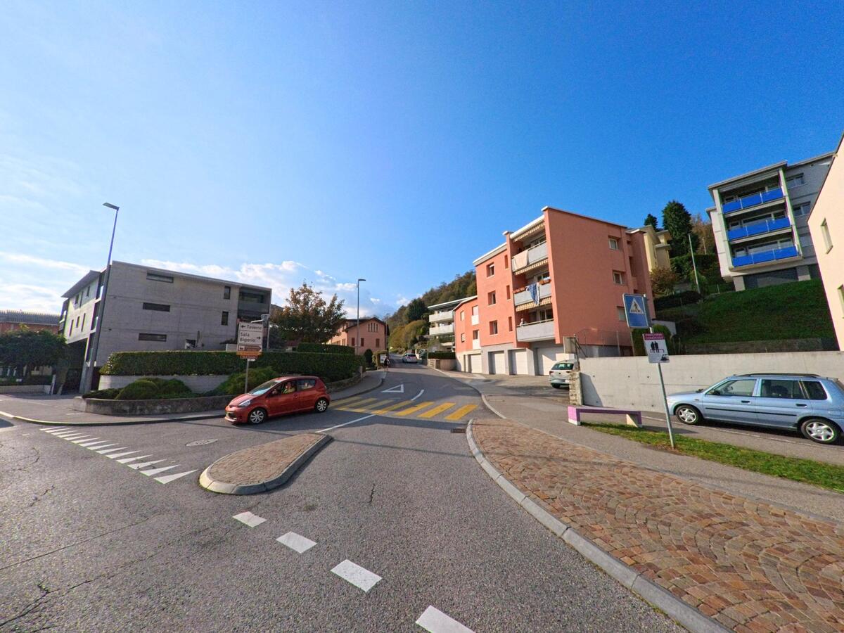Eine Strassenkreuzung mit Zebrastreifen und Mehrfamilienhäusern prägt das Wohngebiet in Capriasca (6950, Kanton Tessin) bei klarem Himmel.