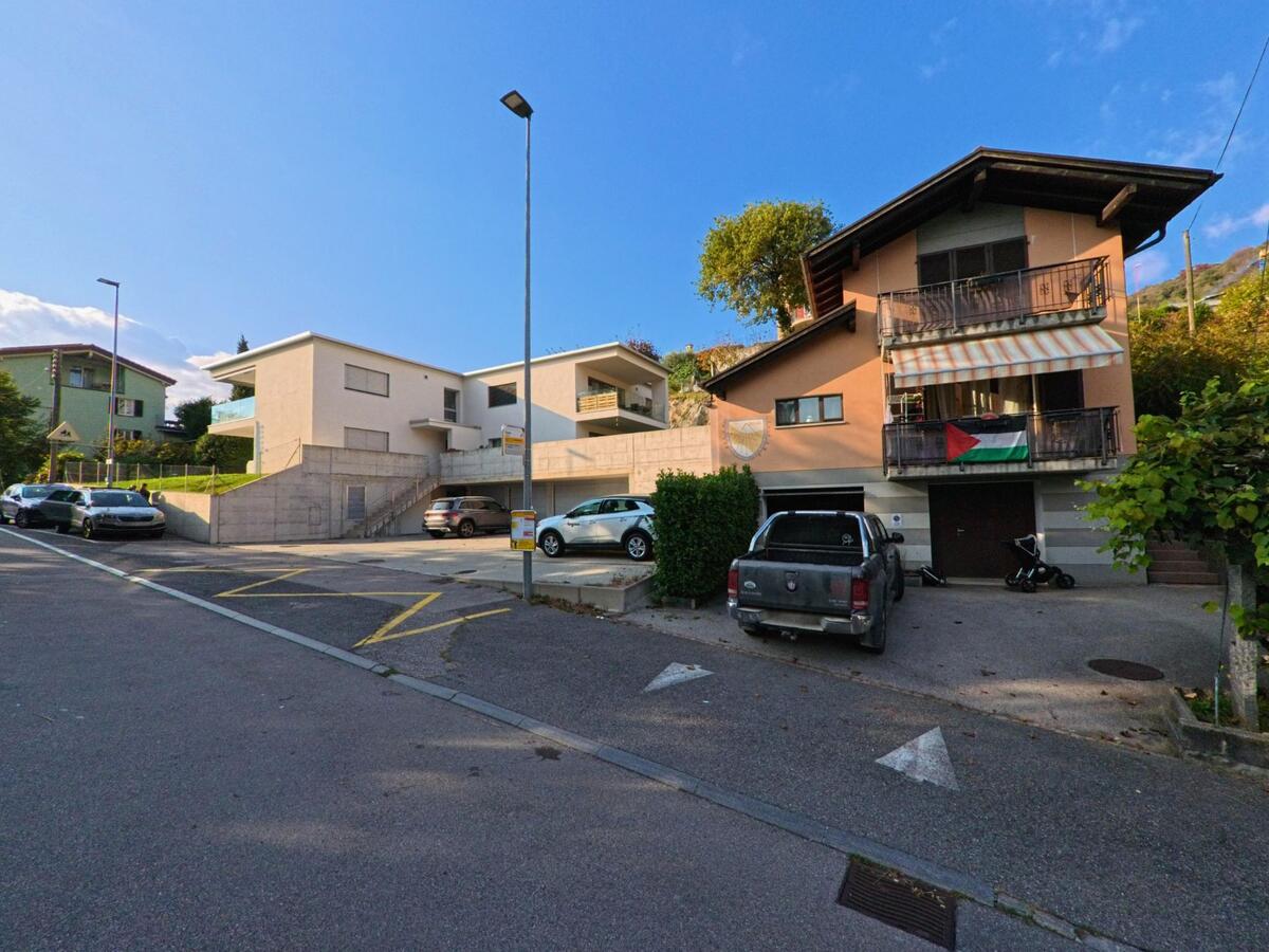 Moderne Wohnbauten und ein älteres Wohnhaus stehen entlang einer Quartierstrasse mit Parkplätzen in Capriasca (6950, Kanton Tessin) unter wolkenlosem Himmel.