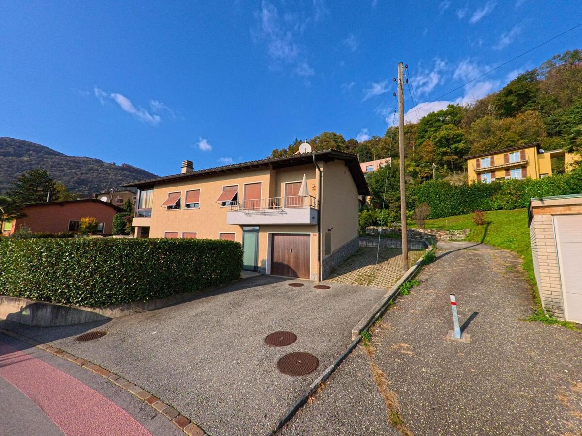 Ein Wohnhaus mit Garage und Balkon liegt an einem schmalen Zufahrtsweg am Hang in Capriasca (6950, Kanton Tessin) vor bewaldeter Umgebung.
