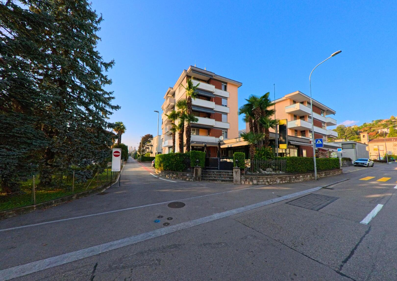 An einer ruhigen Strassenkreuzung in Caslano (6987, Kanton Tessin) stehen moderne Wohngebaeude mit Balkonen und Palmen unter klarem Himmel.
