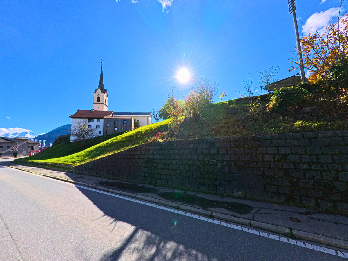 Entlang einer Strasse auf einer Gruenboeschung steht die Kirche mit Blick ueber Cazis (7405, Kanton Graubünden).