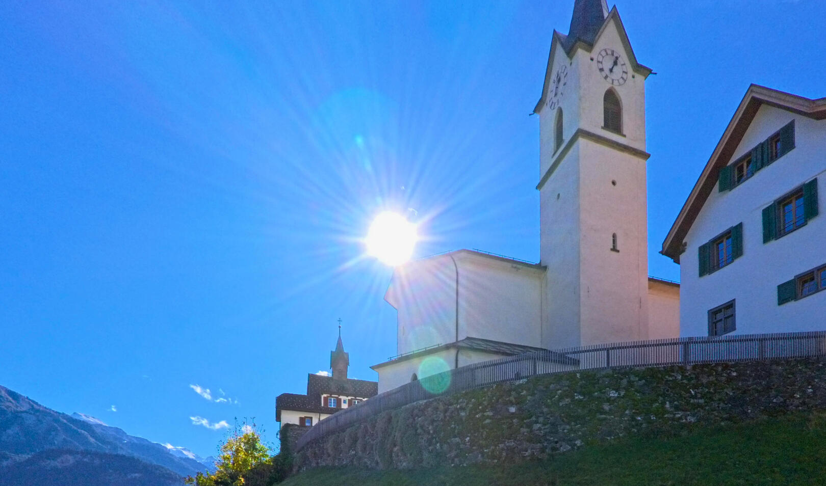 Vor blauem Himmel erhebt sich der Kirchturm in Cazis (7405, Kanton Graubünden), wobei das Sonnenlicht die helle Fassade und die umliegenden Gebäude klar hervorhebt.