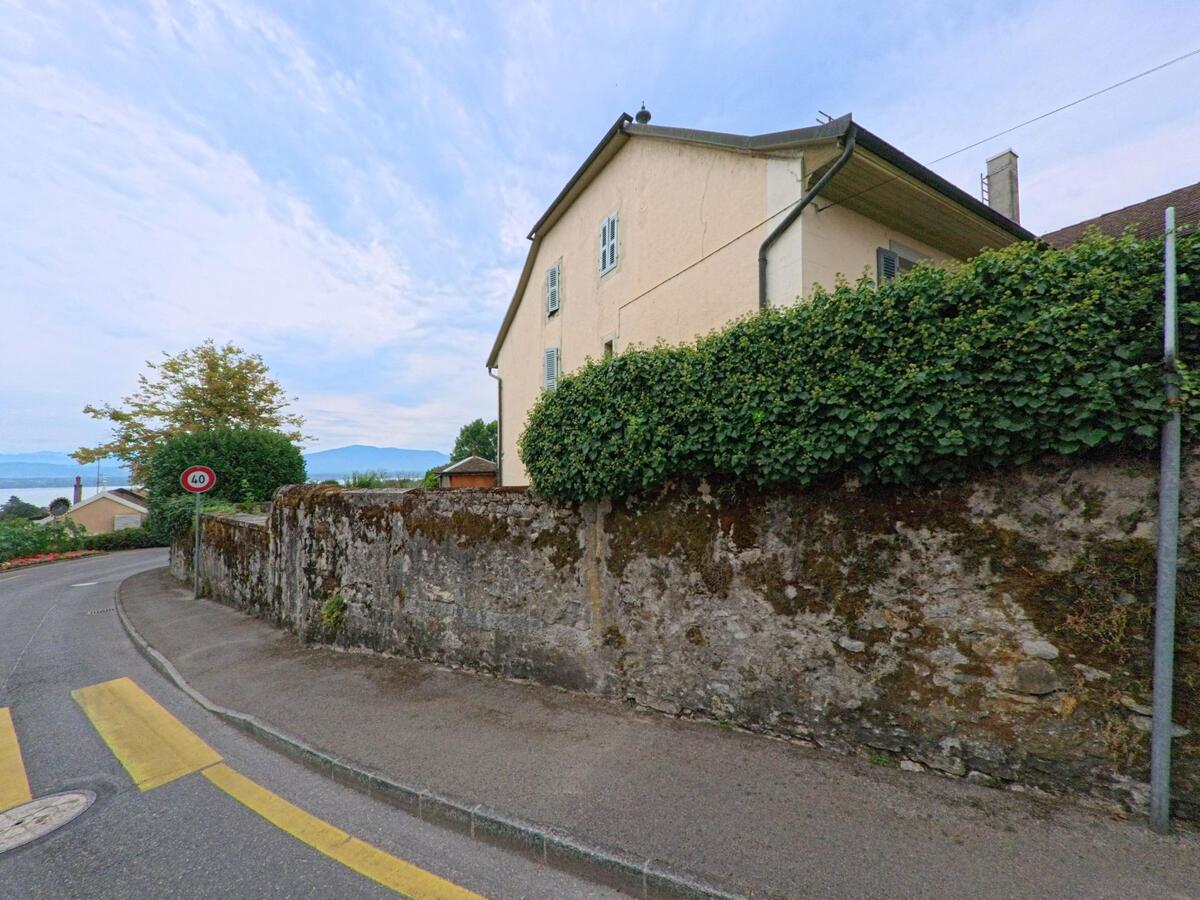Eine Strasse in Celigny (1298, Kanton Genf) fuehrt an einer alten Steinmauer und einem Wohnhaus vorbei, im Hintergrund ist der Genfersee sichtbar.