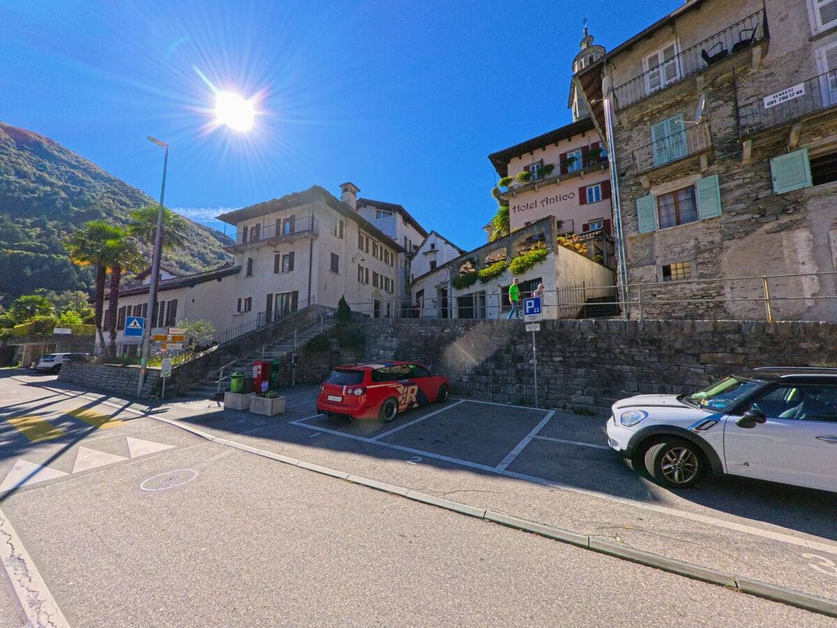 Im Zentrum von Centovalli (6655, Kanton Tessin) stehen traditionelle Steinhäuser und das Hotel Antico unter strahlender Sonne an einer kleinen Strasse mit Parkplätzen.