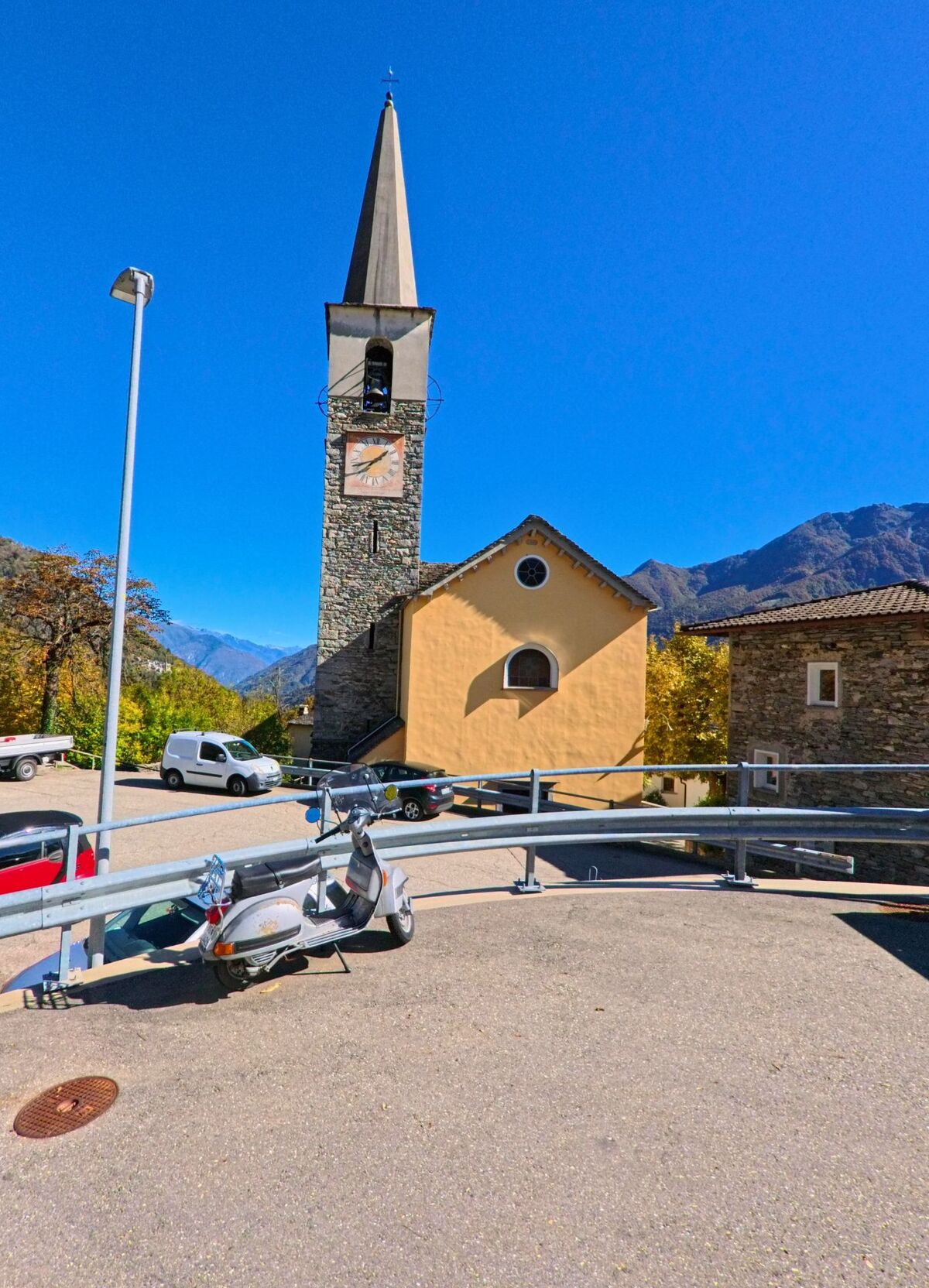 Eine kleine Kirche mit gelber Fassade und hohem Glockenturm aus Stein steht in Centovalli (6658, Kanton Tessin) vor den Bergen, mit geparkten Fahrzeugen im Vordergrund.