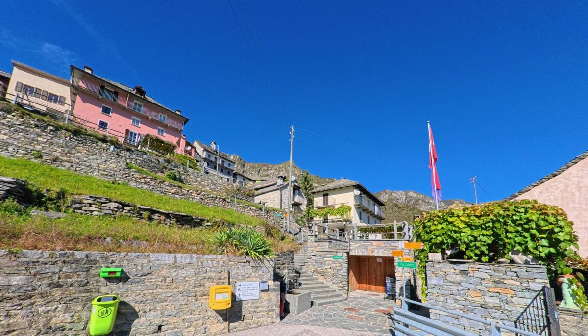 In Centovalli (6658, Kanton Tessin) führen steinerne Treppen durch das terrassenförmig angelegte Dorf mit bunten Häusern und einer Schweizer Flagge vor blauem Himmel.