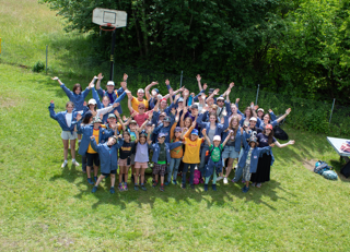 Auf einer Wiese in Richterswil (8805, Kanton Zürich) posiert eine grosse Cevi-Gruppe mit erhobenen Armen vor einem Waldrand für ein gemeinsames Gruppenbild.