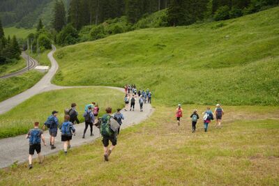 Auf einer gewundenen Bergstrasse in Bubendorf (4416, Kanton Baselland) wandert die Cevi Jungschar Bubendorf als grosse Gruppe durch eine gruene, weite Landschaft.