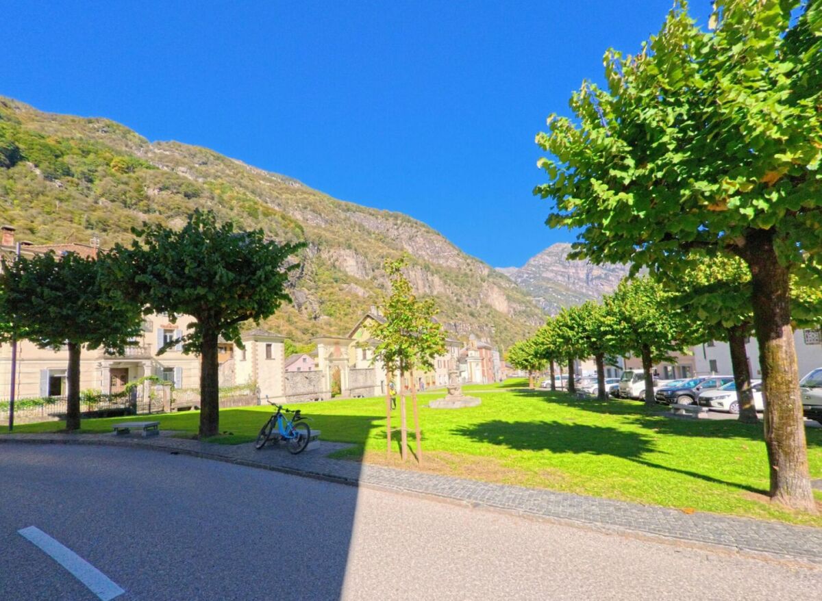 Eine begruente Allee mit Baeumen und Parkflaeche im Zentrum von Cevio (6675, Kanton Tessin) liegt vor einer Kulisse aus Bergen und Hausern.