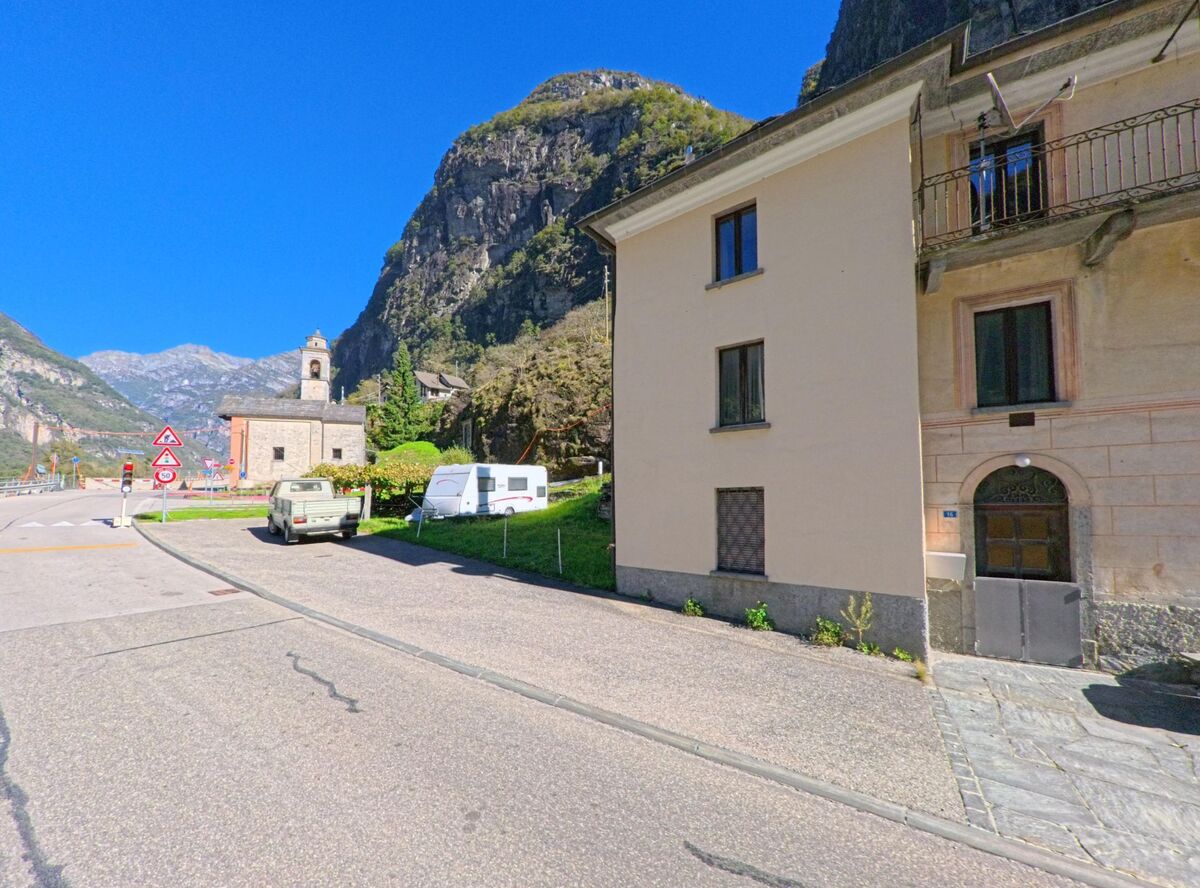 Eine kleine Kirche mit Glockenturm steht am Strassenrand in Cevio (6675, Kanton Tessin), umgeben von Bergen und Wohnhaeusern.