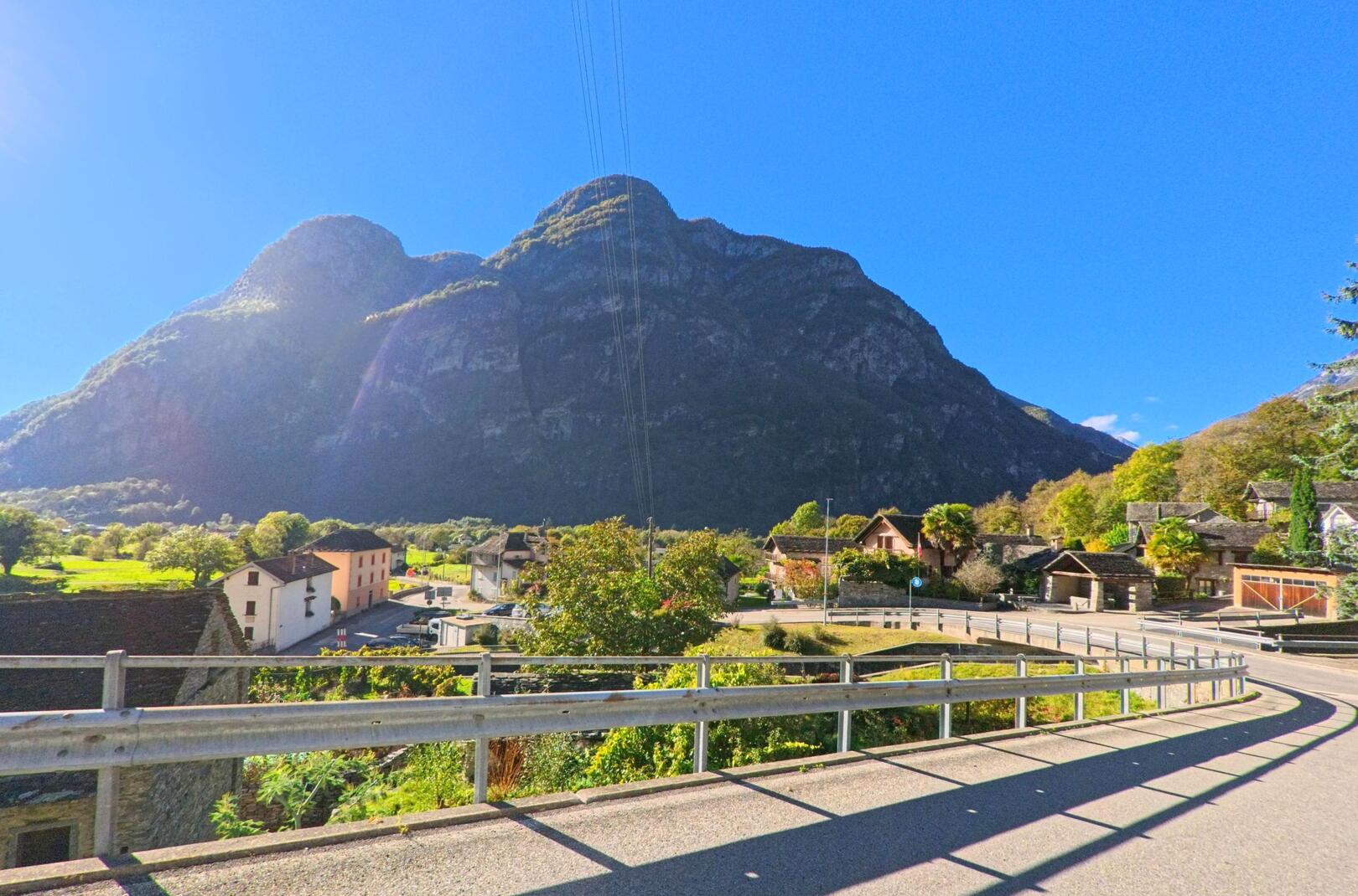 Eine kurvige Strasse in Cevio (6675, Kanton Tessin) führt an traditionellen Steinhäusern vorbei, während im Hintergrund mächtige Berge aufragen