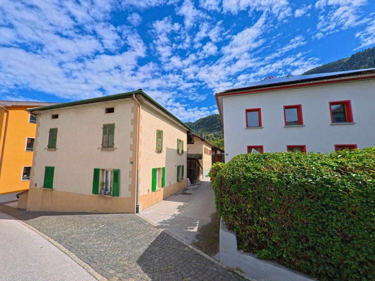 Eine enge Gasse in Chalais (3966, Kanton Wallis) wird von Wohnhaeusern mit gruenen Fensterlaeden und modernen Fassaden gesaeumt.