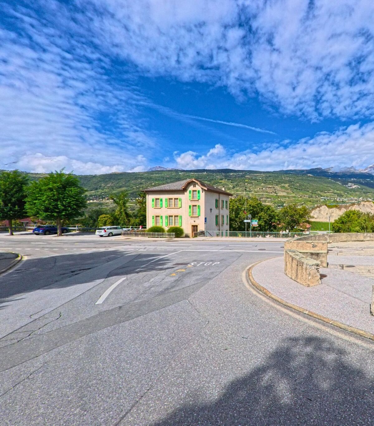 Ein Haus mit grünen Fensterläden steht in Chalais (3966, Kanton Wallis) an einer Strassenkreuzung, im Hintergrund sind Weinberge am Hang sichtbar.