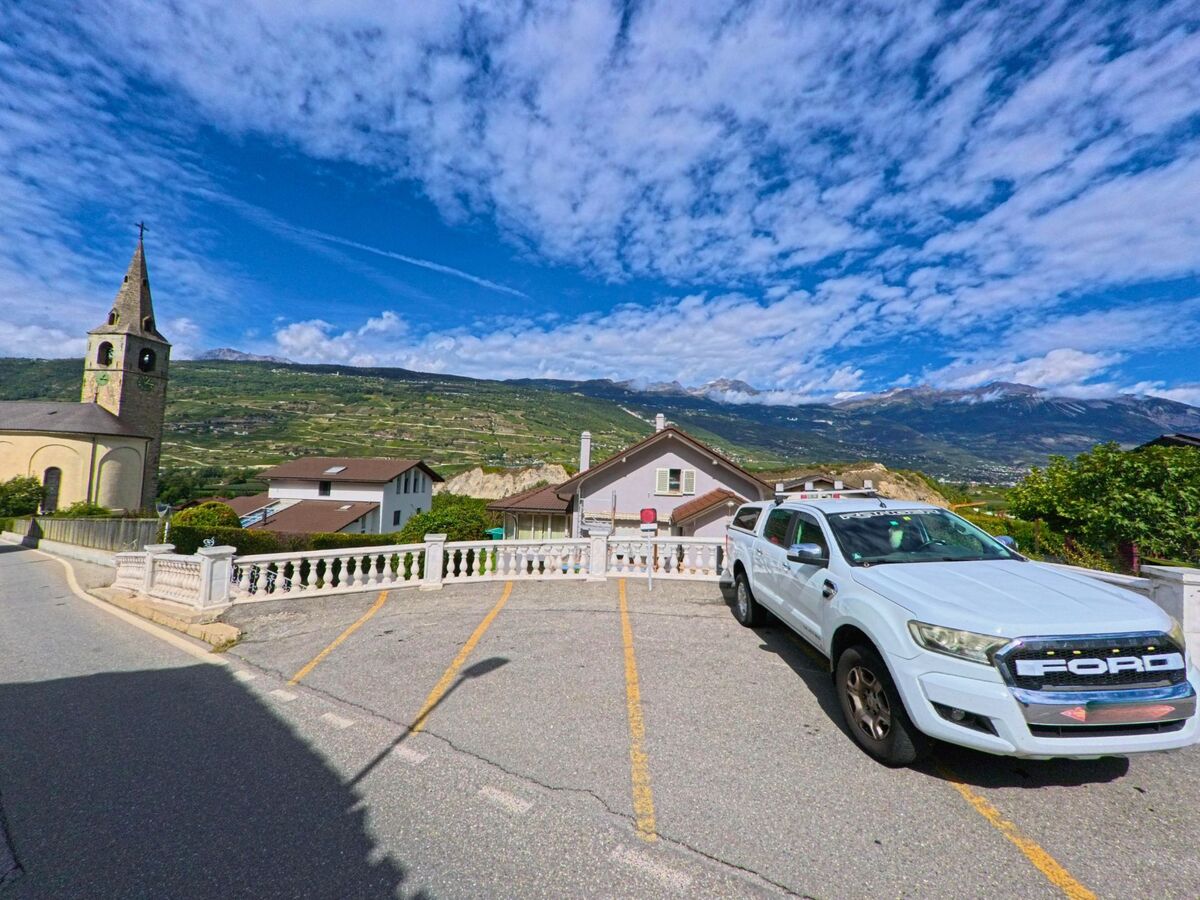 Ein geparkter weisser Wagen steht in Chalais (3966, Kanton Wallis) vor der Kirche, im Hintergrund breiten sich die Weinberge und die Alpen aus.
