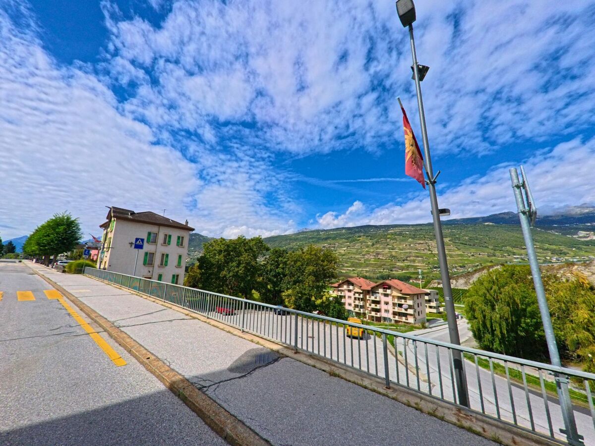 An einer Strasse in Chalais (3966, Kanton Wallis) weht eine Fahne am Laternenmast, im Hintergrund sind Wohnhaeuser und Weinberge am Hang sichtbar.