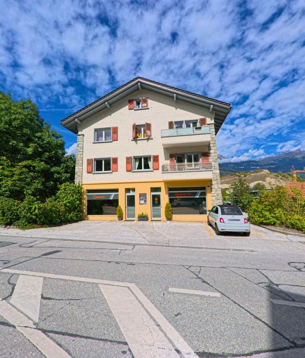 Ein Wohn- und Geschäftshaus in Chalais (3966, Kanton Wallis) steht unter blauem Himmel, vor dem Gebäude befindet sich ein kleiner Parkplatz mit einem Auto.