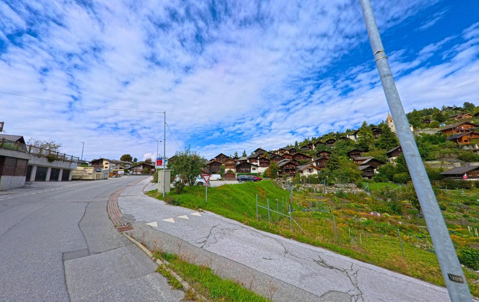 Von der Strasse in Chalais (3967, Kanton Wallis) fällt der Blick auf terrassenförmig angelegte Gärten und eine Hanglage mit traditionellen Holzhäusern.