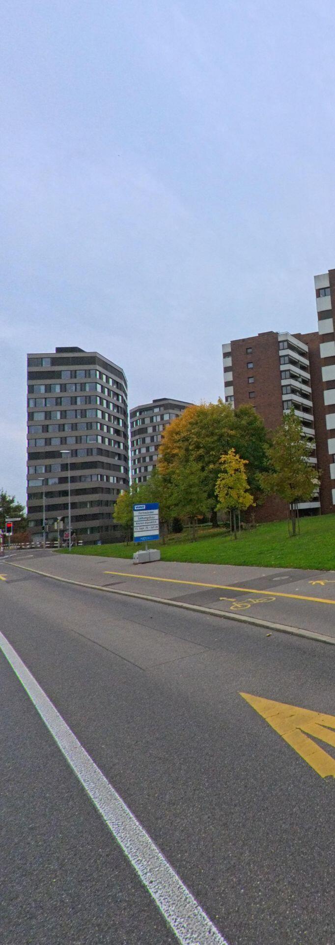 In Cham (6330, Kanton Zug) ragen mehrere hohe Wohngebaeude hinter einer Gruenflaeche auf, waehrend eine breite Strasse mit Markierungen vorbeifuehrt.