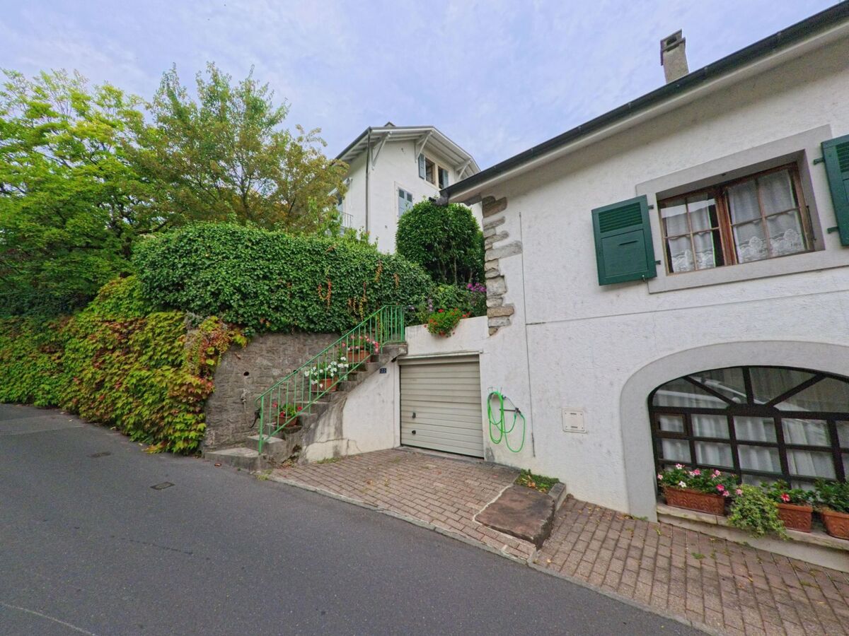 Ein weiss verputztes Haus mit gruenen Fensterlaeden und Garage steht an einer engen Strasse in Chambesy (1292, Kanton Genf).