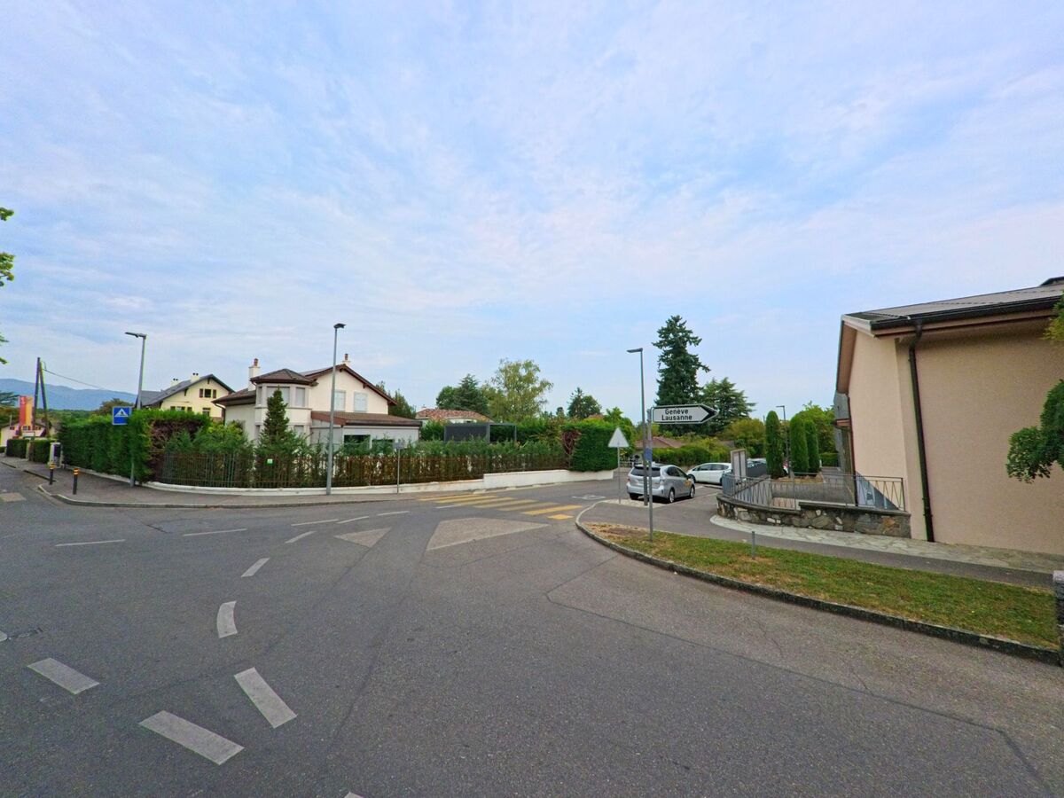 An einer Kreuzung in Chambesy (1292, Kanton Genf) stehen Wegweiser in Richtung Genf und Lausanne, umgeben von Wohnhaeusern und Hecken.