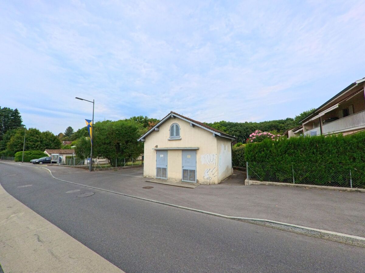Ein kleines, helles Gebäude mit verschlossenen Türen steht an einer Strasse in Chambesy (1292, Kanton Genf), umgeben von Wohnhäusern und Bäumen.