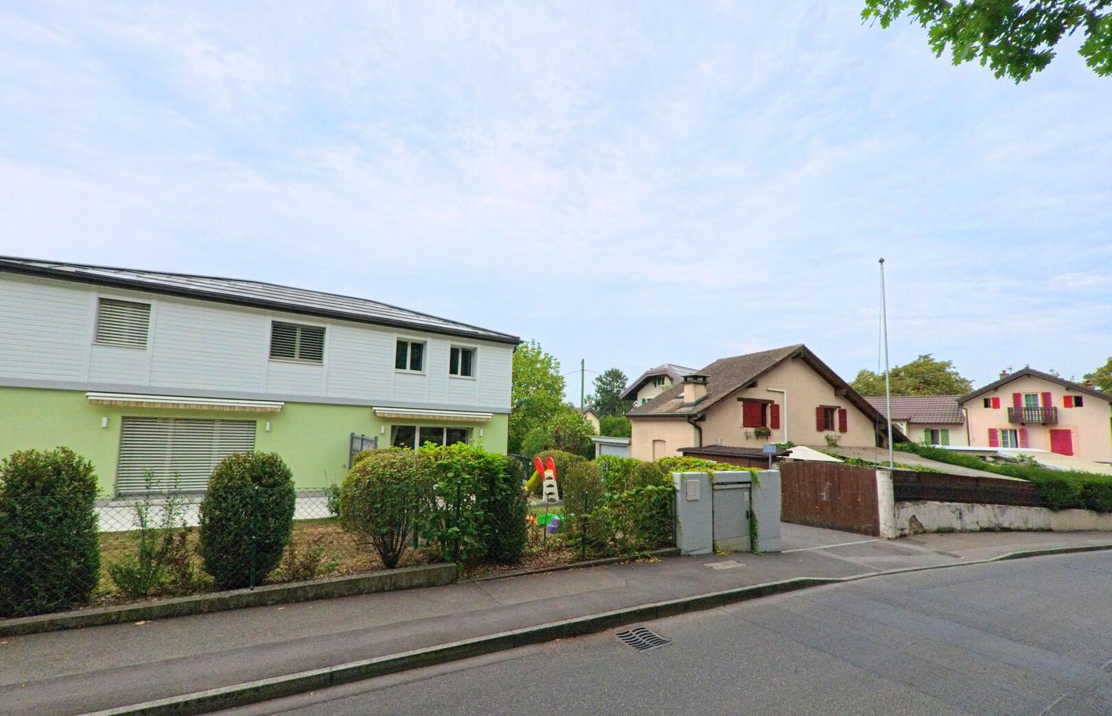 Mehrere Wohnhaeuser mit roten Fensterlaeden und moderner Fassade stehen entlang einer ruhigen Strasse in Chambesy (1292, Kanton Genf).